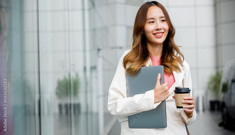 Business woman wearing white suit jacket and smile while go to work outdoor mirror building background. Confident Businesswoman with a cup of coffee and holding Laptop walking outside office building Business woman wearing white suit jacket and smile while go to work outdoor mirror building background. Confident Businesswoman with a cup of coffee and holding Laptop walking outside office building