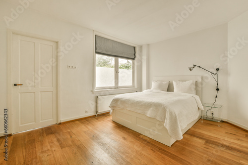 a bedroom with wood floors and white walls  including a large bed in the corner of the room on the right is a window