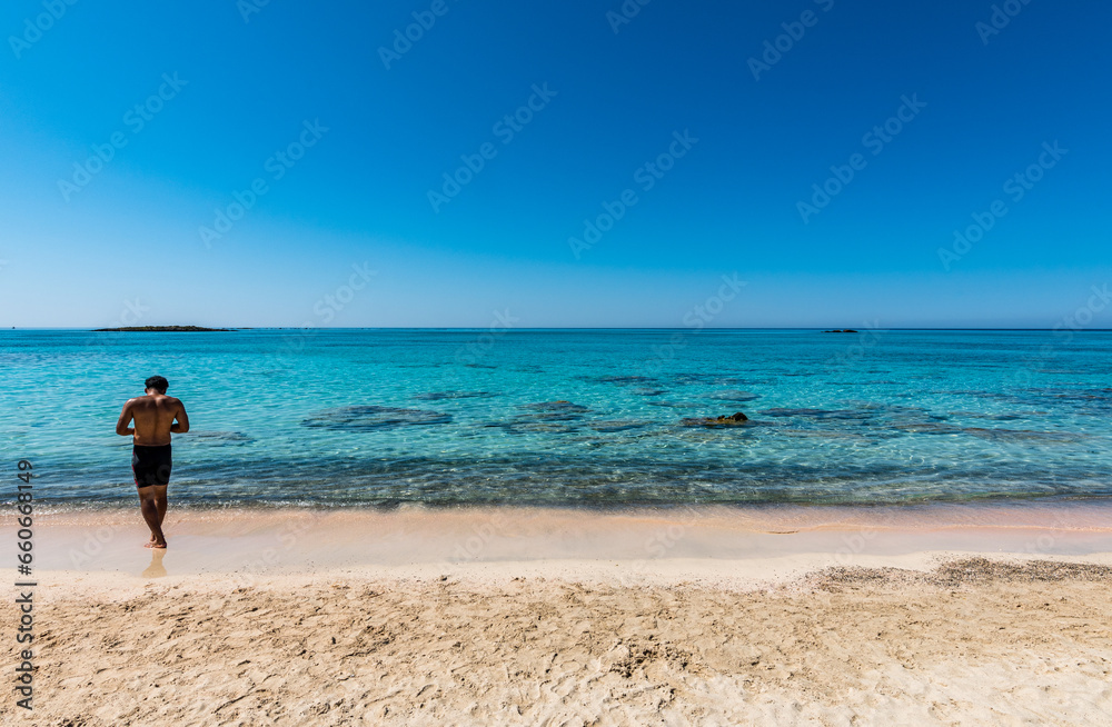 Black boy by the sea. Spectacular panorama of Elafonissi Beach in Crete with Turquoise Water and the famous pink sand. The man is wearing a swimsuit and is sunny-side up.