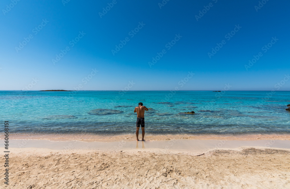 Black boy by the sea. Spectacular panorama of Elafonissi Beach in Crete with Turquoise Water and the famous pink sand. The man is wearing a swimsuit and is sunny-side up. Black boy by the sea. Spectacular panorama of Elafonissi Beach in Crete with Turquoise Water and the famous pink sand. The man is wearing a swimsuit and is sunny-side up.