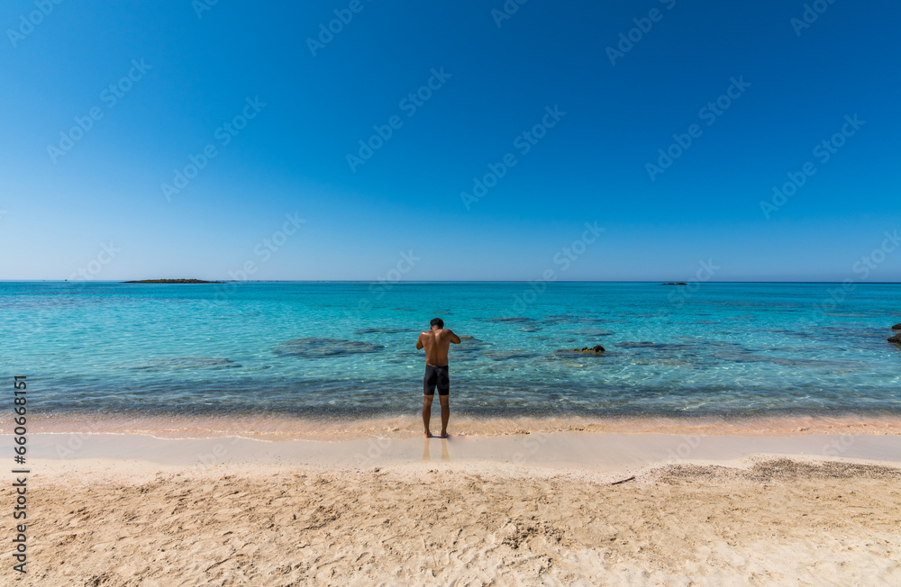 Black boy by the sea. Spectacular panorama of Elafonissi Beach in Crete with Turquoise Water and the famous pink sand. The man is wearing a swimsuit and is sunny-side up.