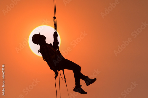 Silhouette of a construction worker hanging on a safety harness against an orange sky at sunset, Thailand photo