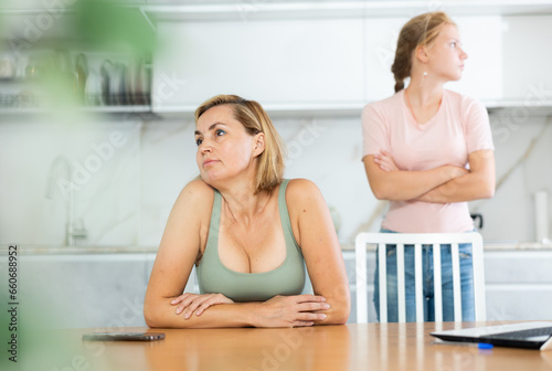 Portrait of upset woman sitting in home kitchen on background of offended frustrated teenage daughter standing behind. Family conflicts and communication problems concept
