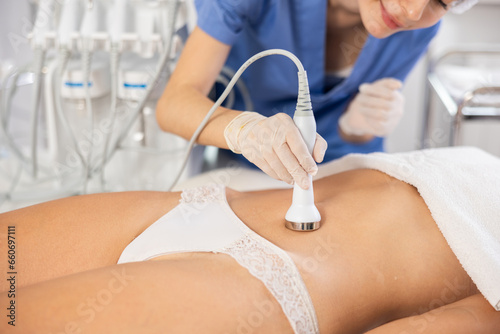 Female patient undergoing ultrasonic cavitation procedure on belly at modern aesthetic medicine clinic to reduce cellulite and adipose fat, improve body shape and reduce circumference