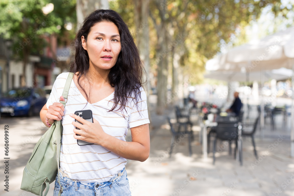 Asian woman with shoulder bag and smartphone in hand walking through city streets.