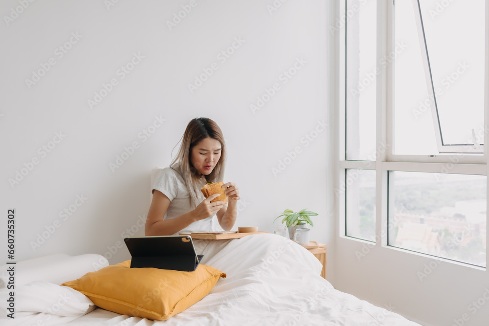 Asian Thai woman having breakfast, eating homemade sandwich while working at in apartment, using tablet in the morning time, busy at home. Asian Thai woman having breakfast, eating homemade sandwich while working at in apartment, using tablet in the morning time, busy at home.
