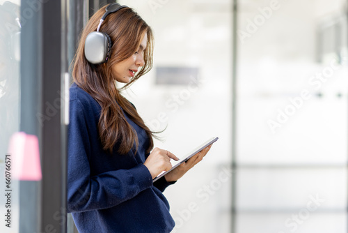 Young adult happy smiling Hispanic Asian student wearing headphones standing indoor using digital tablet in university campus or at virtual office. College female student learning remotely.