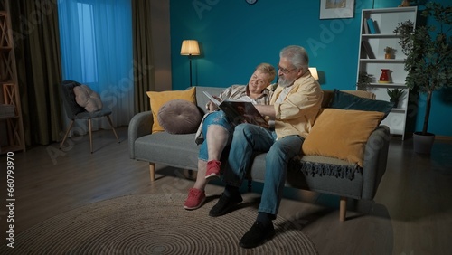In the frame, an elderly couple sits on a couch in an apartment, against a blue wall. They are looking at, leafing through a photo album. They are chatting, reminiscing about old times, nostalgic