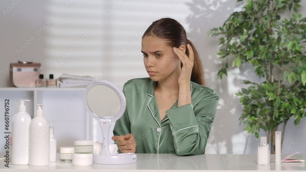 A woman with problematic facial skin looks disappointedly in the mirror. A woman without makeup and wearing green silk pajamas sits at her dressing table in the morning. The concept of problem skin. A woman with problematic facial skin looks disappointedly in the mirror. A woman without makeup and wearing green silk pajamas sits at her dressing table in the morning. The concept of problem skin.