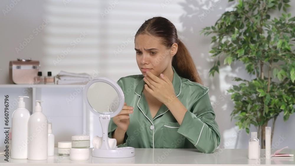 A woman with problematic facial skin looks disappointedly in the mirror. A woman without makeup and wearing green silk pajamas sits at her dressing table in the morning. The concept of problem skin. A woman with problematic facial skin looks disappointedly in the mirror. A woman without makeup and wearing green silk pajamas sits at her dressing table in the morning. The concept of problem skin.