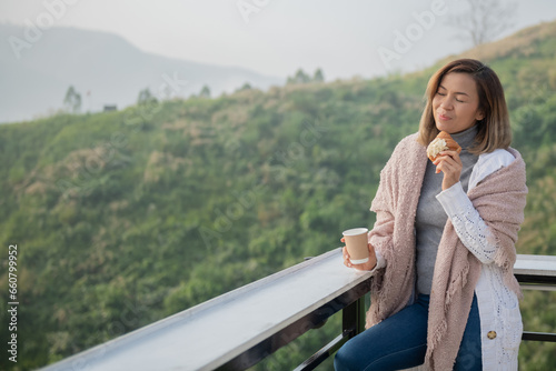 Relaxed woman breathing fresh air holding coffee cup and croissant for breakfast in autumn in a forest sitting on balcony and looking at mountains and green nature.Trekking concept..