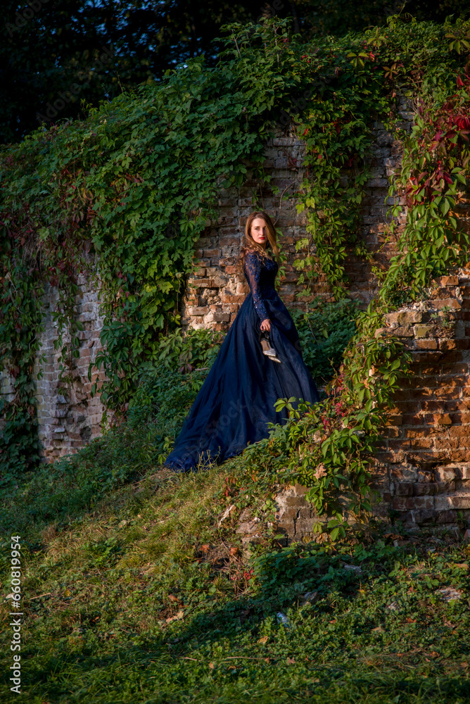 Beautiful brunette woman with curly hair and red lips in long blue dress posing near brick wall in forest Beautiful brunette woman with curly hair and red lips in long blue dress posing near brick wall in forest