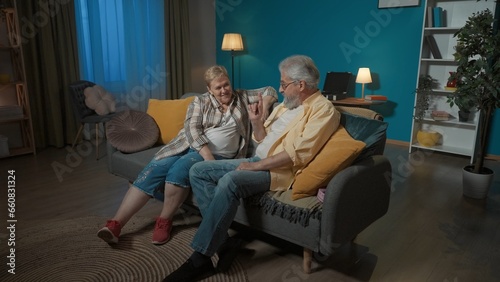 In the picture, an elderly couple is sitting on a sofa in a room. The man is sitting next to a hidden pink box under a pillow. A gift for a woman, wants to surprise her, she does not see or know
