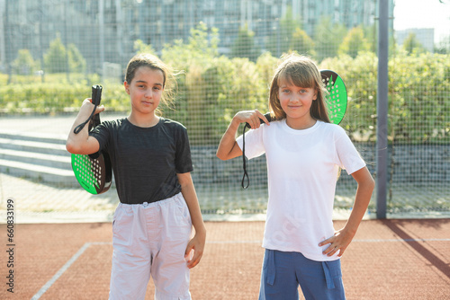 Kids and sports concept. Portrait of smiling girls posing outdoor on padel court with rackets and tennis balls