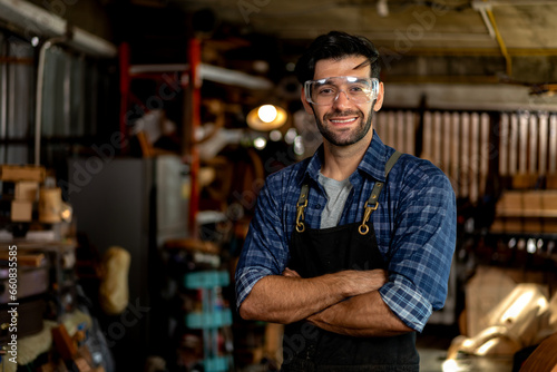 Portrait of a young handsome carpenter wearing a plaid shirt and apron, smiling in his workshop.