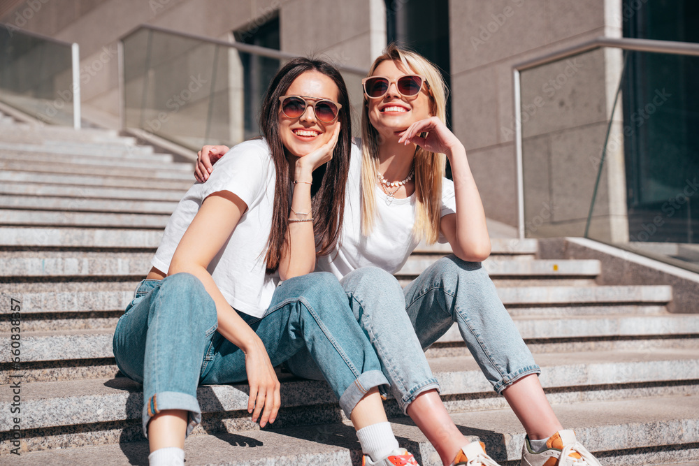 Two young beautiful smiling hipster female in trendy summer white t-shirt and jeans clothes. Carefree women posing in the street. Positive models having fun outdoors. Cheerful and happy. In sunglasses