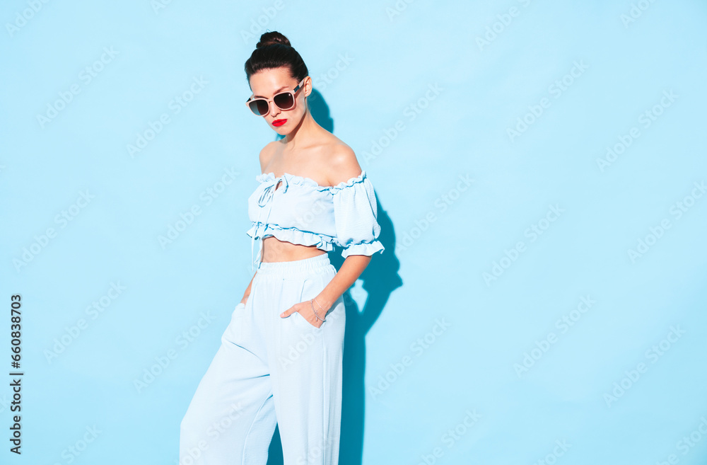 Young beautiful confident female in trendy summer clothes. Sexy carefree woman posing near blue wall in studio. Hot brunette model having with red lips. Cheerful and happy. Isolated. In sunglasses Young beautiful confident female in trendy summer clothes. Sexy carefree woman posing near blue wall in studio. Hot brunette model having with red lips. Cheerful and happy. Isolated. In sunglasses