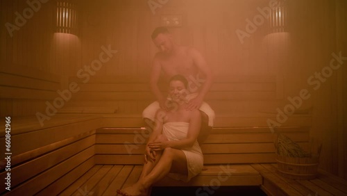A married couple spends time in a Finnish sauna. The sauna is filled with steam. The man and the women are covered with towels. He massages her shoulders while she tells him something and smiles.