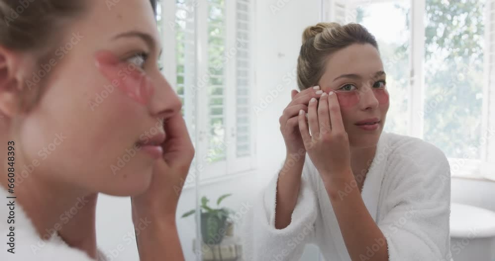 Happy biracial woman applying under-eye patches looking in mirror in bathroom, slow motion