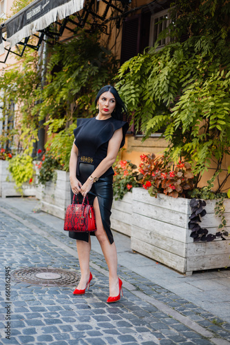 Full length portrait of confident business lady wearing black blouse, skirt and red high heels standing on cobblestone city street with trendy bag in hands. Caucasian woman thoughtfully looking aside.