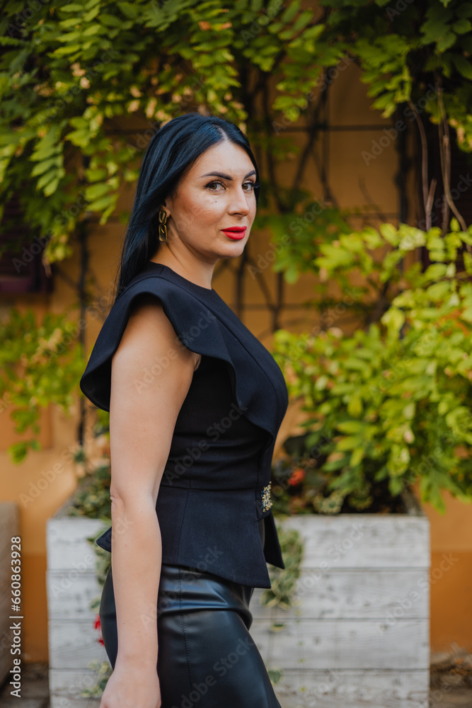 Side view of successful business woman dressed in elegant black blouse and leather skirt strolling on city street near old buildings with green climbing plants. People and lifestyles concept. Side view of successful business woman dressed in elegant black blouse and leather skirt strolling on city street near old buildings with green climbing plants. People and lifestyles concept.