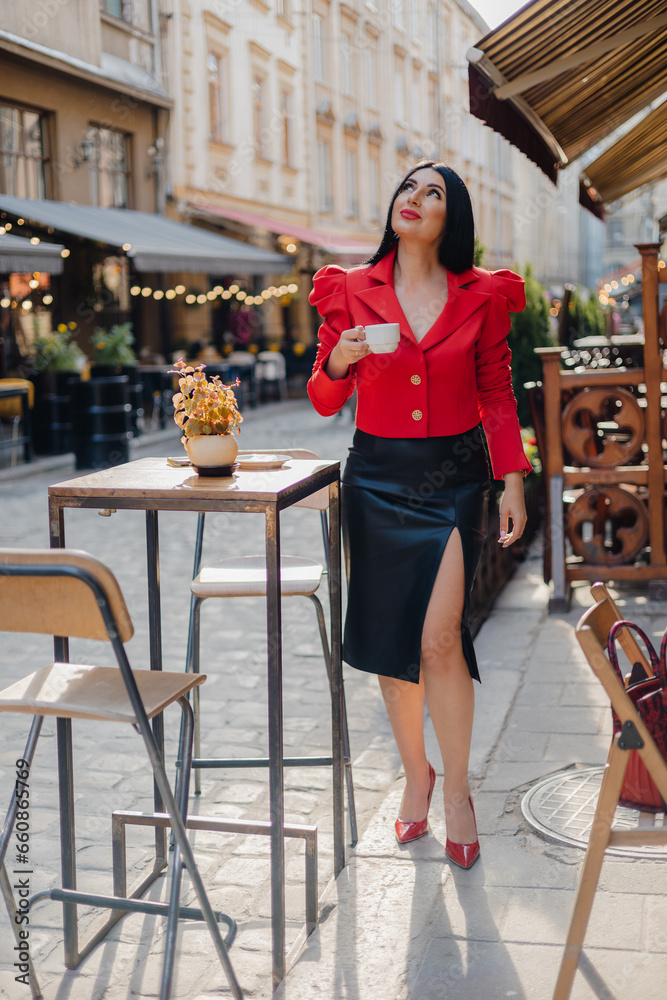 Beautiful female dressed in elegant leather skirt and blouse standing outdoors and looking up against background of ancient houses. Successful businesswoman enjoying aromatic coffee in street cafe.