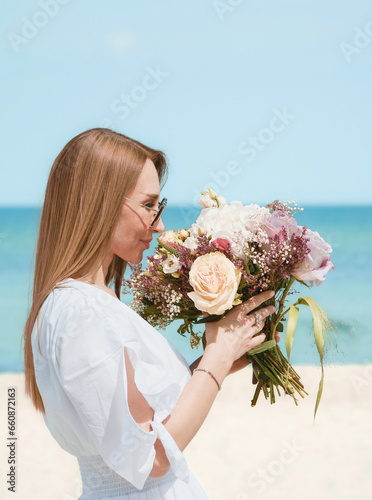 Joyful Young Woman Holding a Bouquet of Flowers on a Beach with Blue Sky Background