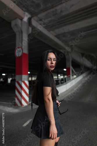 Urban beautiful fashion young woman model in stylish black clothes with a top and skirt walking through a parking lot on the street