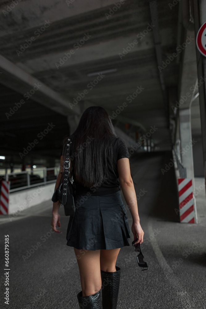 Fashionable beautiful urban woman with long hair in a black top with a skirt walks in a parking lot in the city