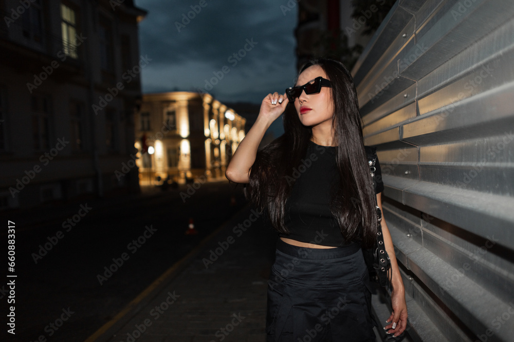 Cool fashionable beautiful Asian party woman in fashion urban outfit puts on stylish sunglasses and walks in the city at night