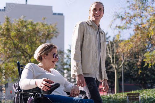 portrait of a woman using electric wheelchair with a young man talking happy as they walk through the city, concept of friendship and diversity photo