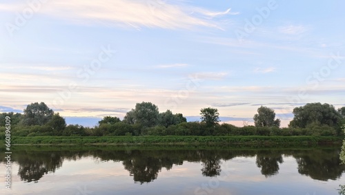 Fototapeta Naklejka Na Ścianę i Meble -  On a quiet summer evening, after sunset, the sky with feathery clouds over the river turns pink and is reflected in the calm water. Shrubs and trees grow on the far grassy bank