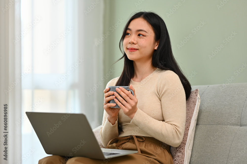 Pretty young woman holding coffee cup and looking away while sitting on couch with laptop Pretty young woman holding coffee cup and looking away while sitting on couch with laptop