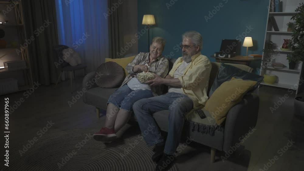 In the shot, an elderly couple are sitting on a couch watching a movie or show on TV and eating popcorn. They are laughing, giggling apparently watching a comedy. HDR BT2020 HLG Material In the shot, an elderly couple are sitting on a couch watching a movie or show on TV and eating popcorn. They are laughing, giggling apparently watching a comedy. HDR BT2020 HLG Material