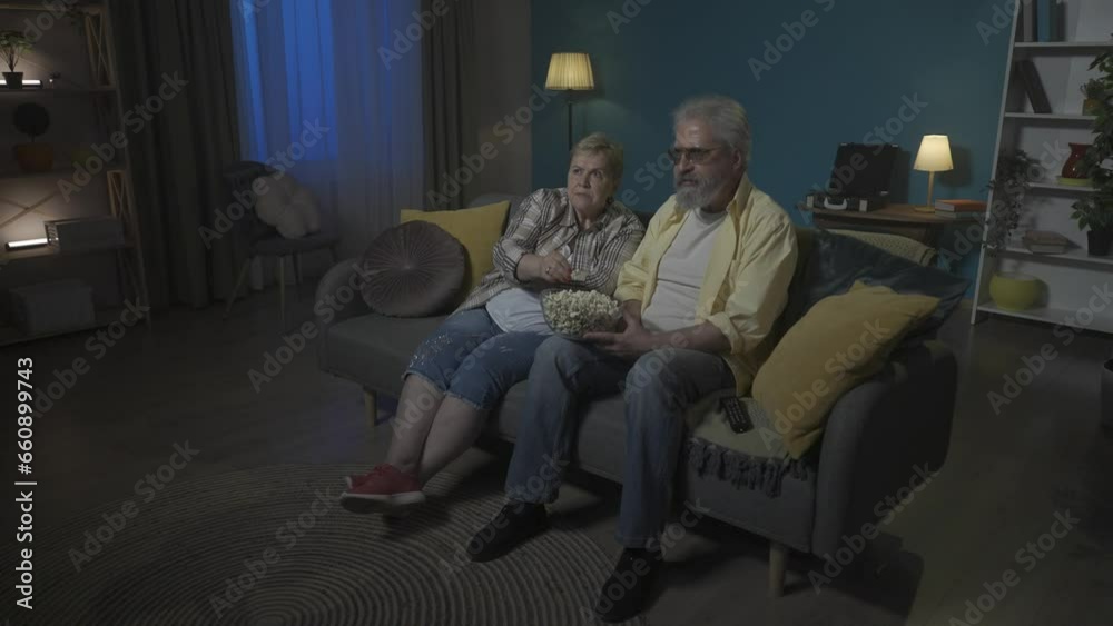 An elderly couple are watching a horror movie on TV and eating popcorn. They are watching carefully, concentrated. The woman is scared and covers her face with her hands. HDR BT2020 HLG Material. An elderly couple are watching a horror movie on TV and eating popcorn. They are watching carefully, concentrated. The woman is scared and covers her face with her hands. HDR BT2020 HLG Material.