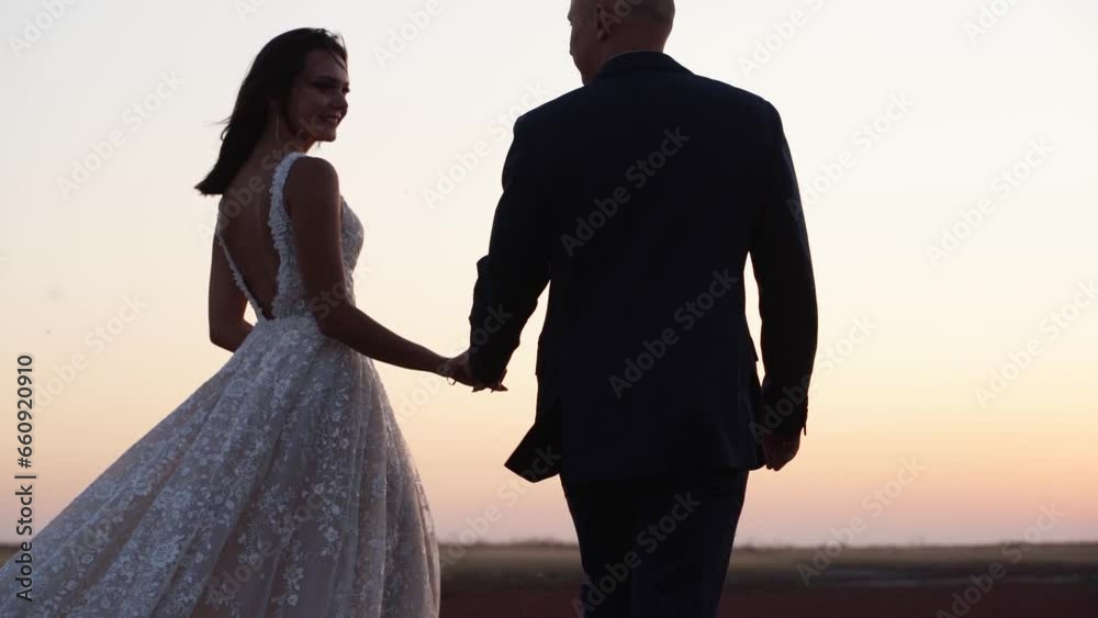 Newlywed couple holding hands, walking across the field at amazing summer sunset. Just married man, woman go on the nature outside at sunrise. Happy wife and husband posing at wedding photoshoot.