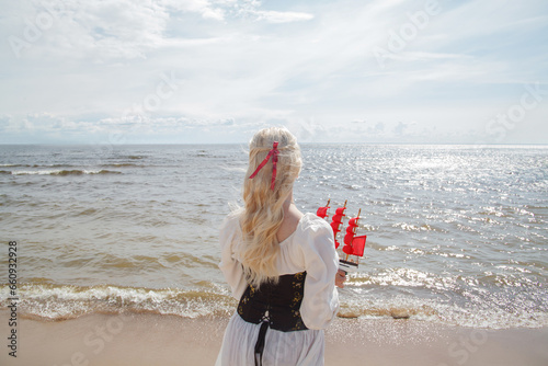 Young romantic blonde woman standing on sea beach with red sails small boat, outdoor portrait photo