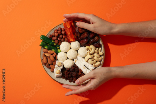 Plate with nuts and sweets in hands on orange background, top view