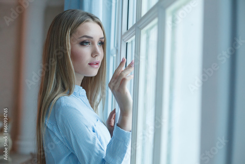 Beautiful woman opening blue curtains looking out window at home