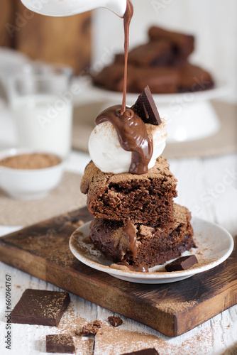 Brownie with a scoop of ice cream cut into pieces and topped with chocolate from a gravy boat. In the background is a northerly table with milk and the rest of the brownies.