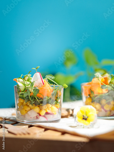 Bowl salad of corn, fresh vegetables and micro-greens in a glass cup. On a wooden table. Blue background, selective focus.