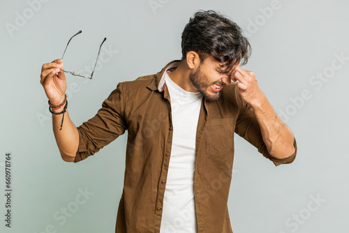Exhausted tired Indian young man takes off glasses, feels eyes pain, being overwork burnout from long hours working. Sleepy exhausted Arabian guy rubbing eyes isolated on studio gray background