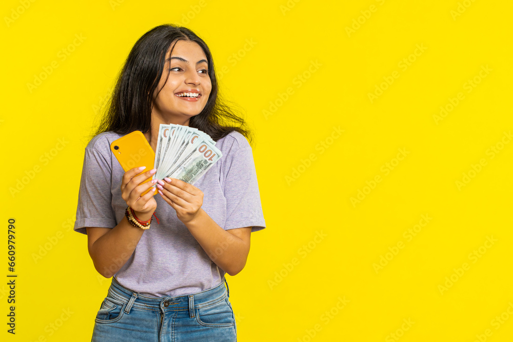 Indian woman looking smartphone display sincerely rejoicing win, receiving money dollar cash banknotes celebrating success victory play lottery game. Lucky rich Arabian girl on yellow background Indian woman looking smartphone display sincerely rejoicing win, receiving money dollar cash banknotes celebrating success victory play lottery game. Lucky rich Arabian girl on yellow background