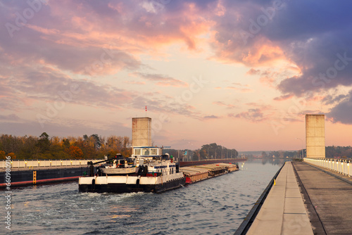 Quadro su tela Scenic view od big cargo freight tugboat barge boat ship sailing by german inland canal Magdeburg bridge against warm sunset sky backrgound