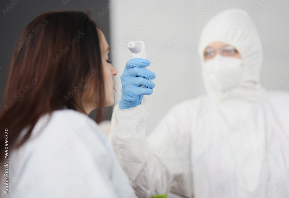 Doctor in protective suit and gloves measures temperature of person. Temperature measurement with non-contact thermometer in the coronavirus pandemic concept