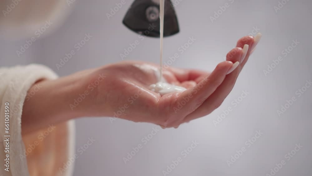 Person pours liquid conditioner for hair into palm in shower. Creamy hair conditioner drips onto female hand in spacious room with blurred background