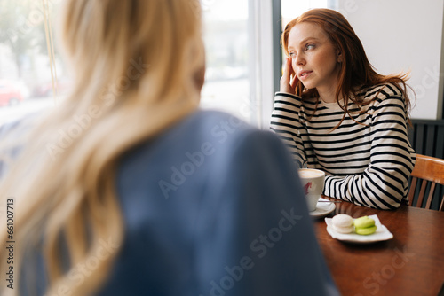 Foto Rear view of unrecognizable blonde female talking to pensive woman friend, spending time together during drinking coffee at cozy cafe, sitting at table by window