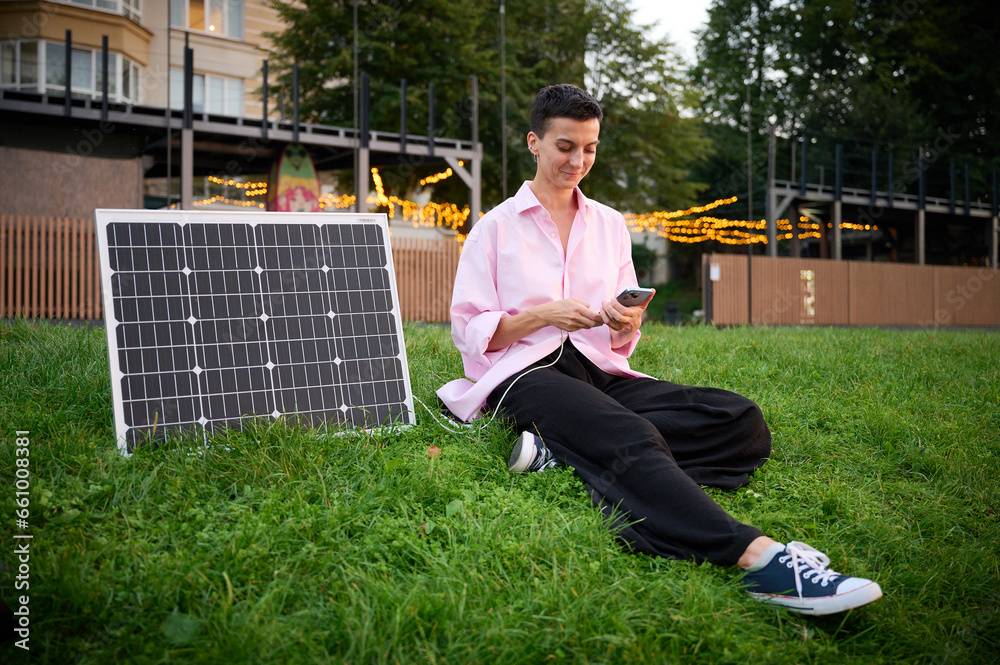 Young woman charging her phone by solar panel. Pretty female in casual clothes sitting on grass and pluging charger in her phone. Renewable energy concept.