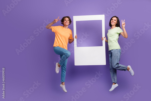 Full body photo of jumping high two sisters showing victory v sign hold window frame zone portrait isolated on violet color background © deagreez