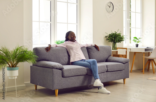 Young girl resting on the couch. Happy introverted African American woman spending weekend at home, sitting on a soft grey sofa in the living room, recharging, relaxing, and enjoying quiet time alone